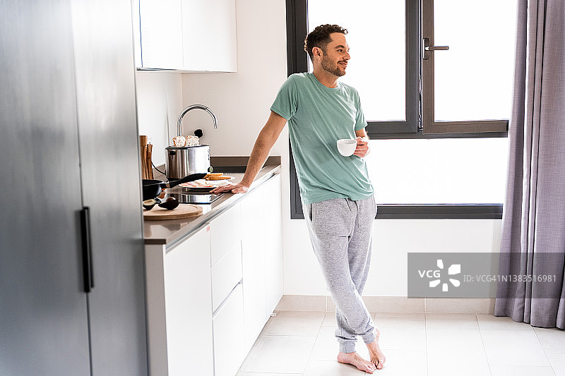 一个男人在家里的厨房里准备早餐时喝着咖啡。图片素材