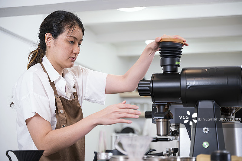 用咖啡的醇厚口感建立顾客忠诚度。女咖啡师用专业的咖啡研磨机研磨咖啡豆来制作咖啡的侧视图。图片素材