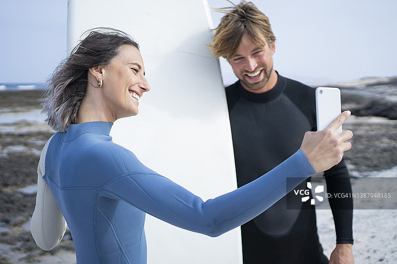 穿着潜水服的幸福夫妇在海滩上用冲浪板自拍图片素材