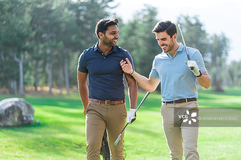 年轻的高尔夫球手们拿着球杆，面带微笑地走在高尔夫球场上，边走边说边笑图片素材