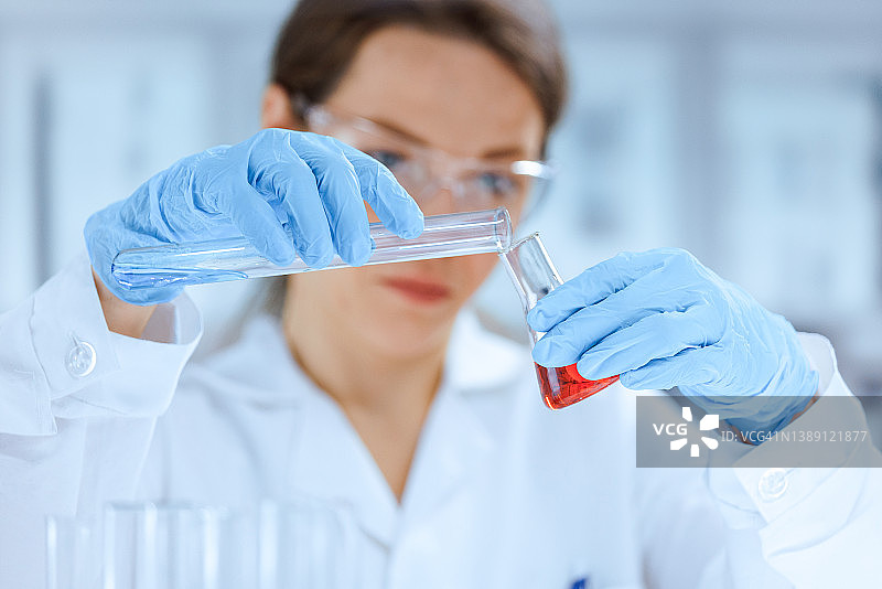一名年轻的白人女化学家穿着实验室的外套，戴着防护手套，在实验室的烧瓶中混合有色液体图片素材