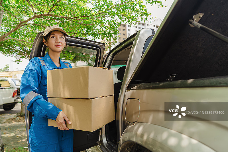 中年亚洲妇女快递员穿蓝色制服，戴棒球帽，从一辆小货车上提着包裹盒图片素材