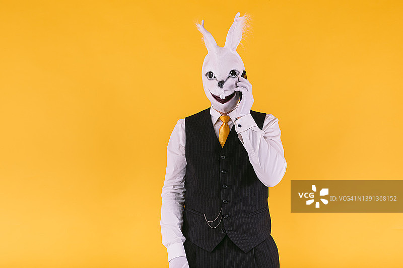 伪装的人戴着兔子面具，穿着西装外套，背心和领带，在黄色的背景上讲着手机。嘉年华，派对，复活节和庆祝的概念。图片素材