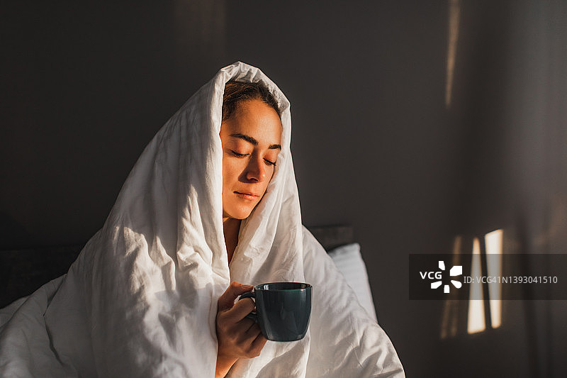 在寒冷的早晨，女人裹着毯子，在床上享受着热咖啡图片素材