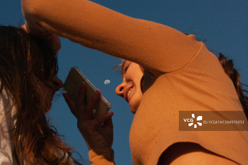 美丽的十几岁的女孩穿着棕色的衣服，用智能手机拍摄女友的特写照片，清澈的蓝天和月亮在背景中图片素材