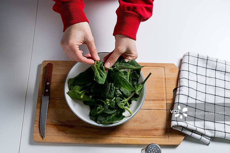 一个女人或十几岁的女孩手里拿着绿色的菠菜叶，背景是一张木制的厨房桌子。在家做饭。素食，纯素食，生食和饮食。农场eco-organic产品。素食主义的概念，纯素食主义，生食。图片素材