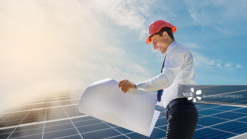 身穿白衬衫、头戴红色安全帽的年轻工程师手持一份项目计划文件，在太阳能发电厂验证光伏板。图片素材