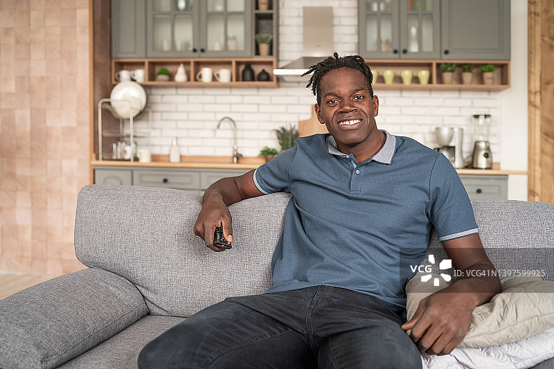 一个迷人的黑人男子的肖像，站在家里，坐在沙发上，拿着一个电视遥控器图片素材