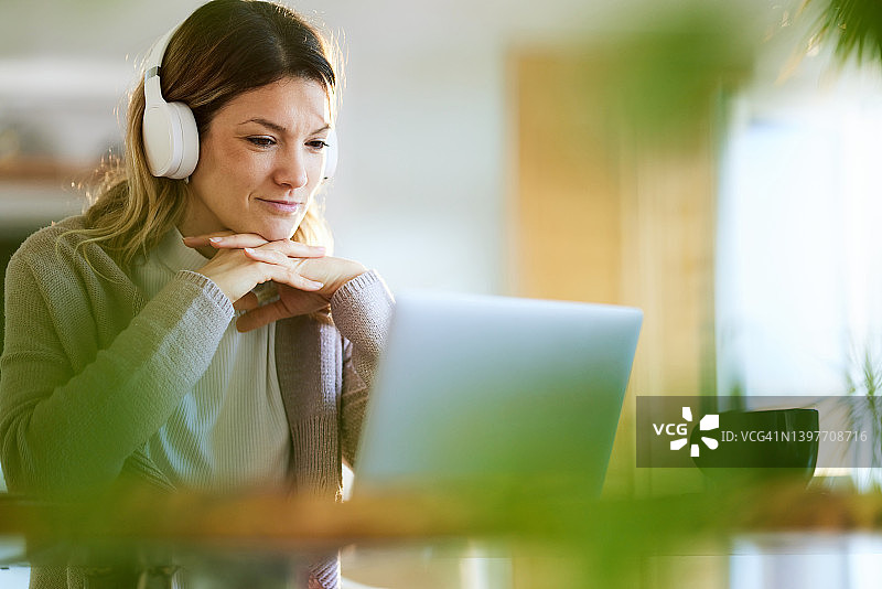 微笑的女人戴着耳机使用笔记本电脑在家办公。图片素材