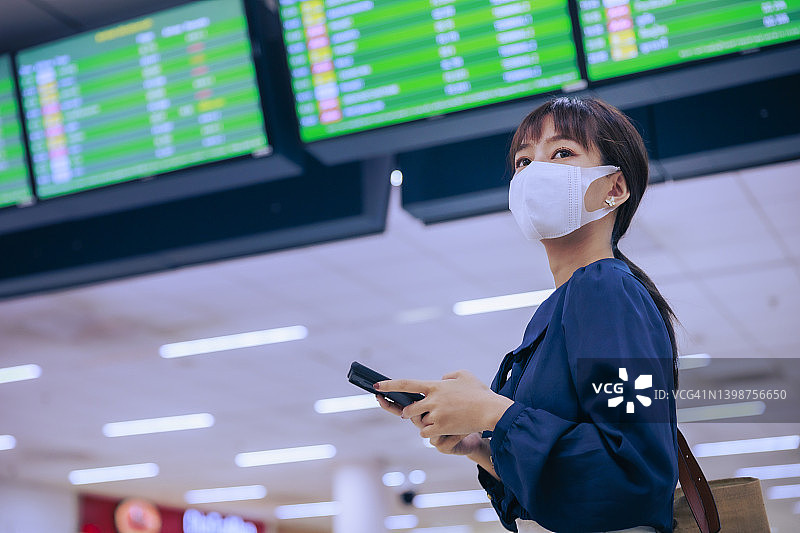 年轻的亚洲女企业家戴着口罩站在机场出境区，用手机阅读电子邮件。商务旅行的概念。图片素材