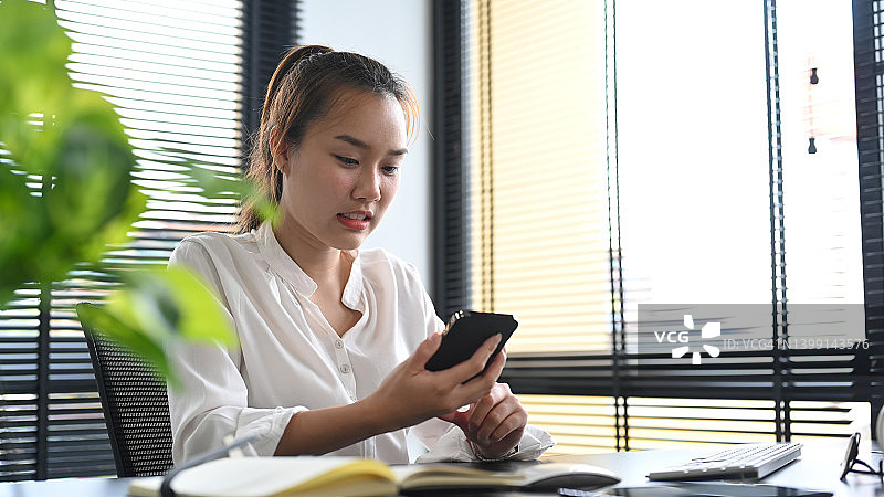 亚洲女性经理坐在工作场所，用手机在社交网络上交流。图片素材