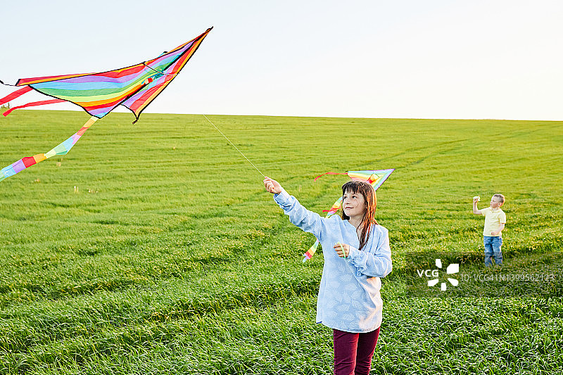 微笑着的女孩和弟弟在高高的草地上奔跑，放着五颜六色的风筝。快乐的童年时光图片素材