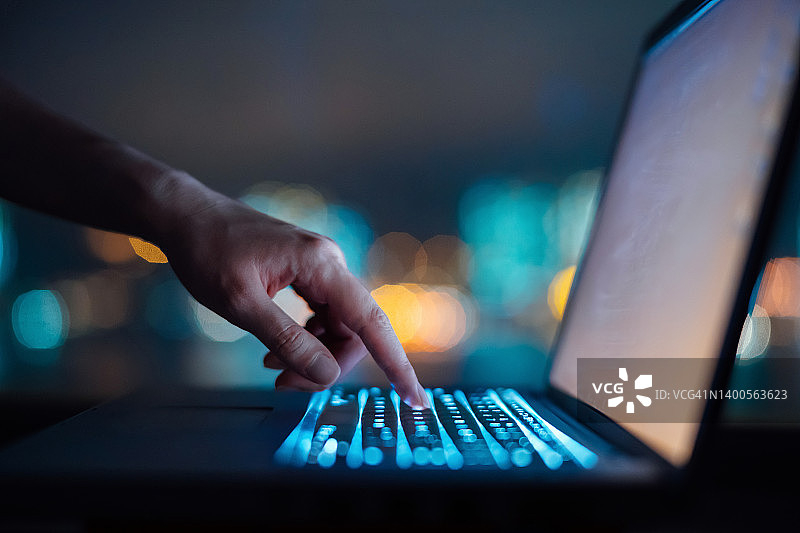 近距离的女人的手在电脑键盘上打字在黑暗中对彩色的散景背景，工作到很晚的笔记本电脑在家里图片素材