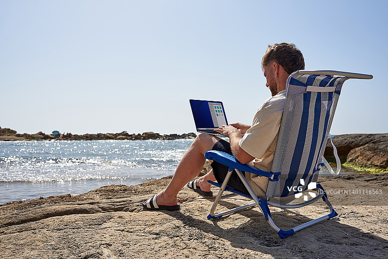 一个年轻人坐在沙滩上的椅子上，用笔记本电脑远程办公图片素材