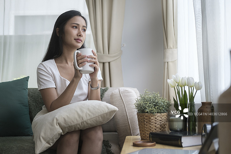 积极的年轻女子坐在舒适的沙发在家里，喝热咖啡在早上。图片素材