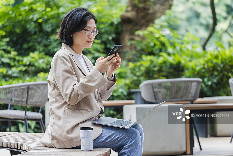 一名亚洲女性在户外咖啡馆一边用笔记本电脑工作一边用手机图片素材