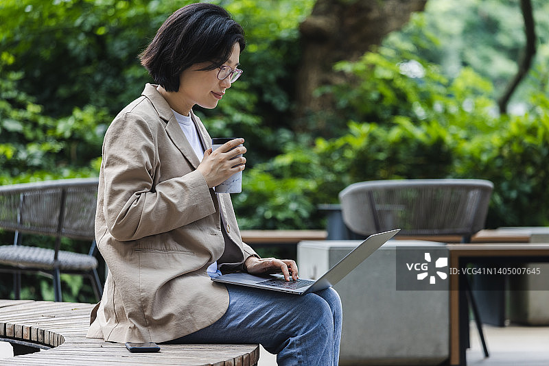一名亚洲女性在户外咖啡馆用可重复使用的咖啡杯喝咖啡时使用笔记本电脑图片素材