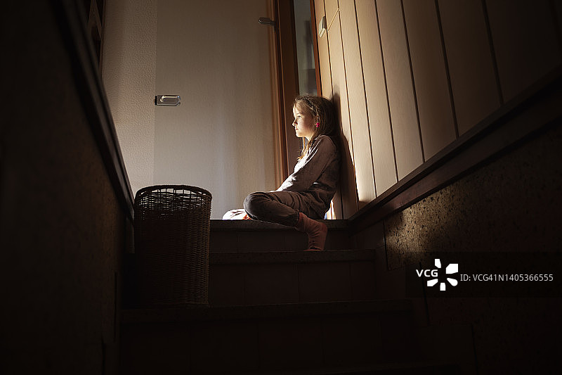 一个孩子坐在通往灯光昏暗的地窖的楼梯顶上的台阶上，面无表情地沉思着。图片素材