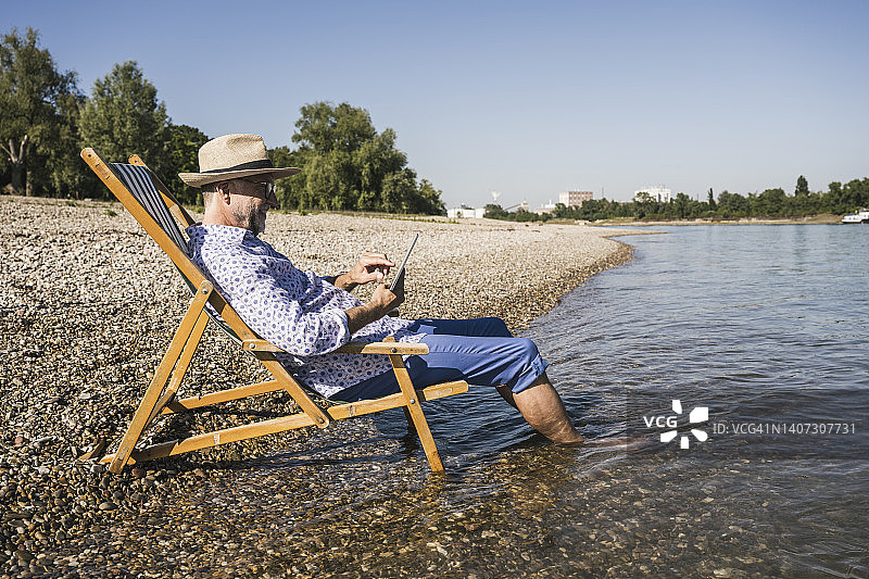 河边的躺椅上，一名微笑的男子正在使用平板电脑图片素材