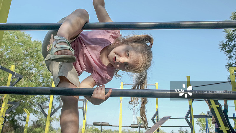小女孩在露天运动场爬体操梯。可爱的小女孩在太阳日爬上城市公园的垂直运动梯。图片素材