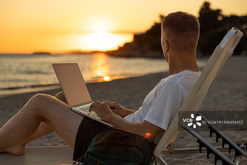 在海滩上欣赏日落的男子，同时在笔记本电脑上工作图片素材
