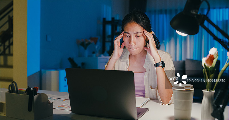 年轻的亚洲女性穿着正装衬衫坐在桌子前与笔记本电脑的屏幕上，认为感到压力，没有想法，使紧急报告会议，加班到午夜在家。图片素材