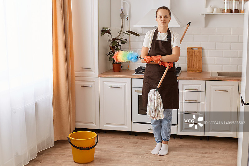 疲惫的家庭主妇在厨房洗地板的全貌，漂亮的年轻女子在做家庭清洁，保持她的公寓整洁，感觉劳累过度，看着镜头。图片素材