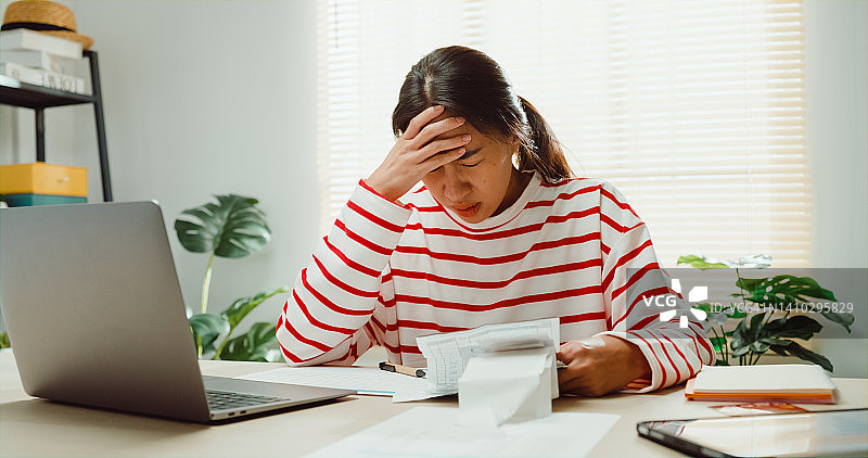 年轻的亚洲女人穿着毛衣坐在桌子前用笔记本电脑检查收据每月的费用账单感觉不快乐压力使用计算器计算债务问题在家里。图片素材