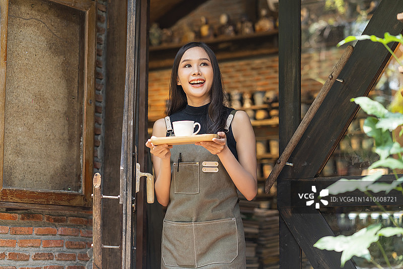 咖啡馆女老板带着积极的幸福微笑着准备为顾客开门，亚洲女店员拿着围裙手捧托盘和咖啡杯站在咖啡店门口热情欢迎顾客图片素材