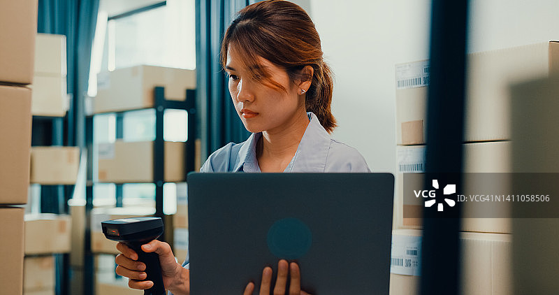 亚洲女商人站在货架前使用条形码机扫描纸盒中的客户数据，注册在线代码，在笔记本电脑上订购明细，以便在仓库快递。图片素材