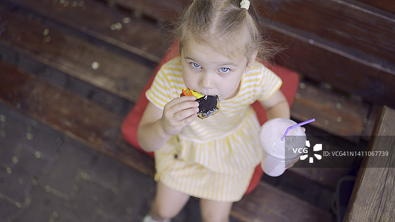 小女孩吃着一个五颜六色的姜饼，手里拿着一杯奶昔。特写可爱的小女孩坐在公园的长凳上，吃饼干和奶昔，看着相机镜头。俯视图图片素材