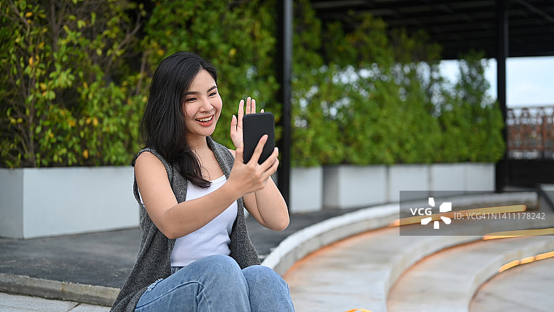阳光明媚的一天，快乐的亚洲女人坐在公园里用手机和朋友视频通话。图片素材