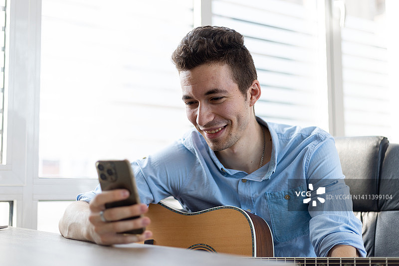 男吉他手坐在室内拿着智能手机，从弦乐器演奏中休息一下，用手机微笑着读着愉快的信息。爱好、消遣、现代无线科技用户概念图片素材