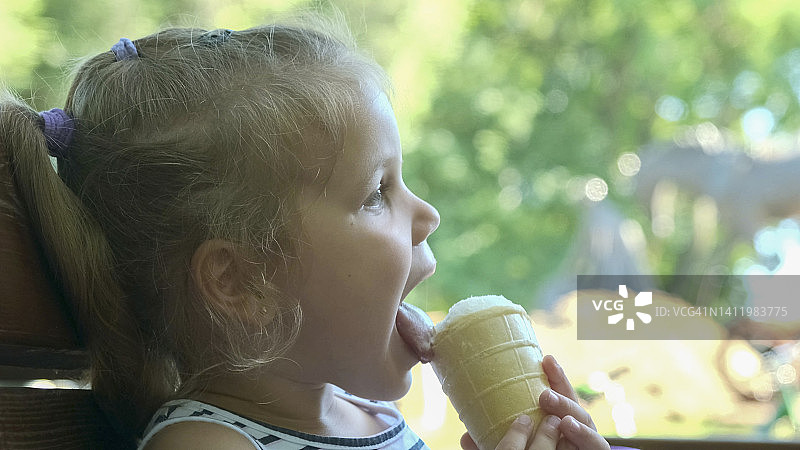 可爱的小女孩在外面吃冰淇淋。特写的金发女孩坐在公园的长凳上吃冰淇淋。图片素材