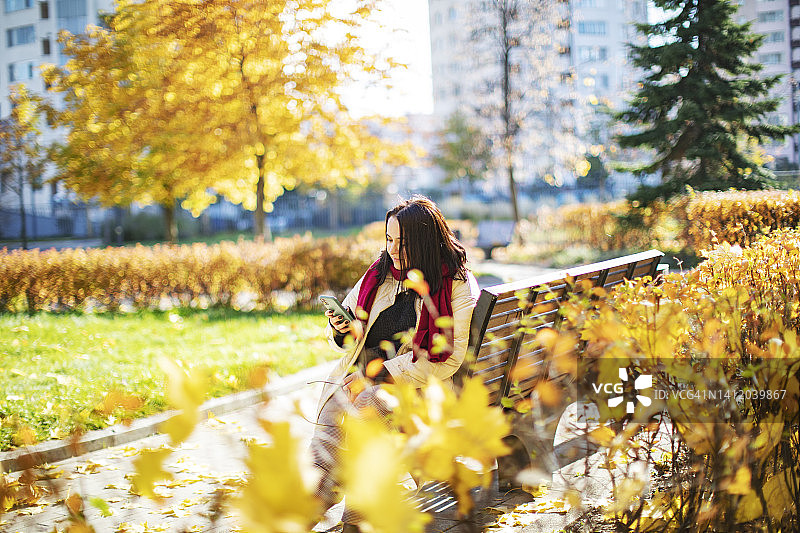年轻的成年女子坐在秋天公园的长凳上使用手机图片素材