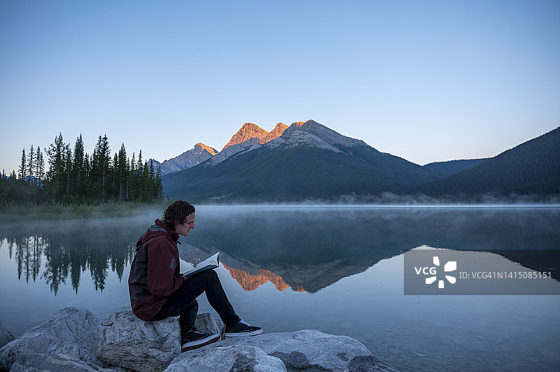在晨光中，一个带着书的年轻人站在湖岸上图片素材