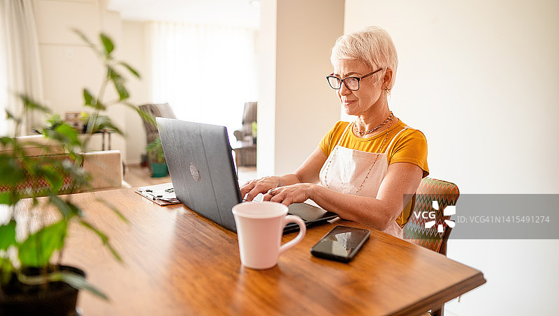 成熟女性坐在家里的桌子前，用笔记本电脑工作图片素材