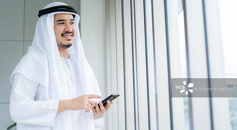 特写肖像阿拉伯商人微笑看向窗外，而持有智能手机在办公室的领导理念图片素材