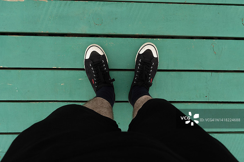 穿着运动鞋的男人的腿。一个男孩或青少年，一个学生站在码头，桥或平台的海洋或湖泊的边缘。木制彩绘墩板色彩鲜艳，临摹空间为文字。一种积极的生活方式的概念，旅游和旅行图片素材