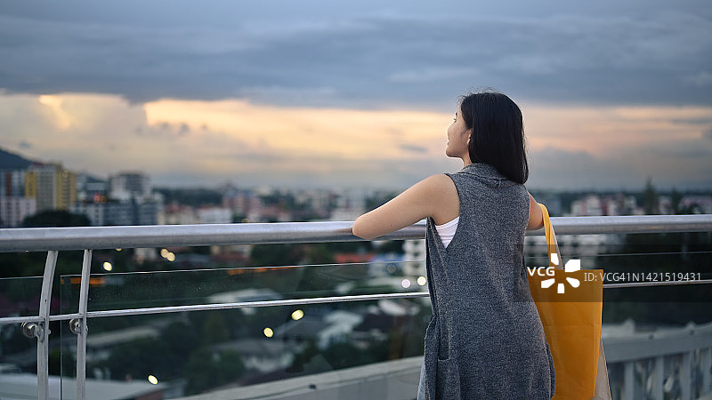时尚的年轻女子站在阳台上俯瞰城市景观和美丽的日落山。图片素材