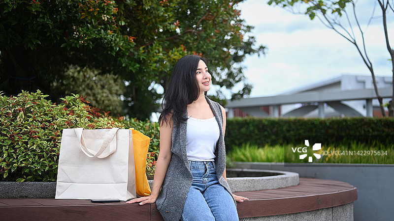迷人的亚洲妇女与她的购物袋坐在美丽的绿色公园的长凳上。图片素材