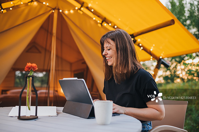 微笑的女性自由职业者使用笔记本电脑在一个阳光灿烂的日子舒适的野营帐篷。豪华户外露营帐篷，适合夏季度假和度假。生活方式的概念图片素材