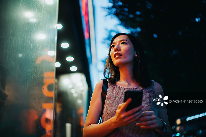 年轻的亚洲妇女一边使用智能手机一边看着时尚精品店外的商店橱窗，走在城市的夜晚，五颜六色的霓虹灯反射在她身上图片素材