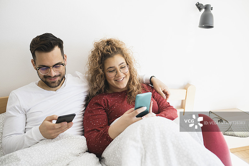 周末的早晨，一对快乐的中年夫妇躺在床上放松，使用智能手机图片素材