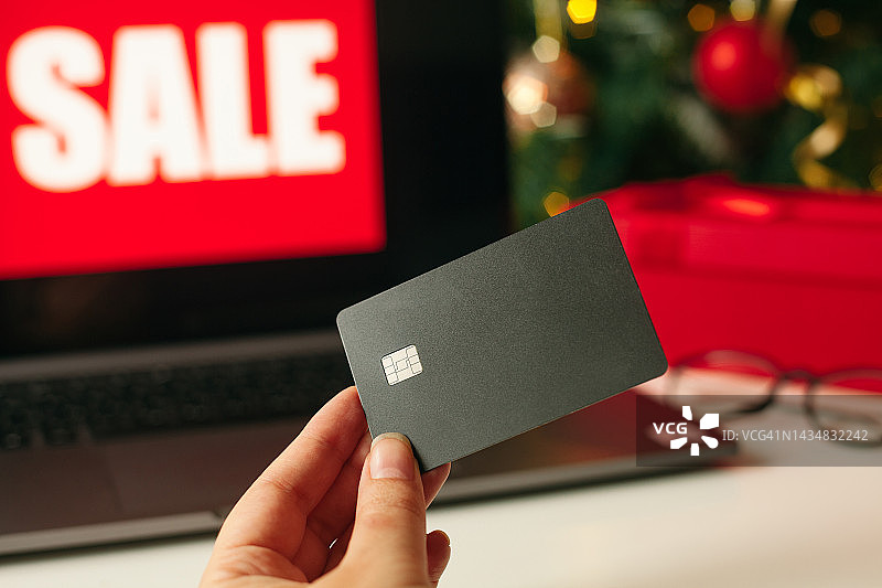 网上购物和圣诞大减价的概念。支付信用卡在手。笔记本电脑屏幕与文字销售，礼品盒，圣诞树背景。图片素材