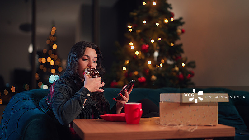 圣诞节期间，年轻女子在家里的客厅里吃着甜食，享受着使用智能手机的乐趣图片素材