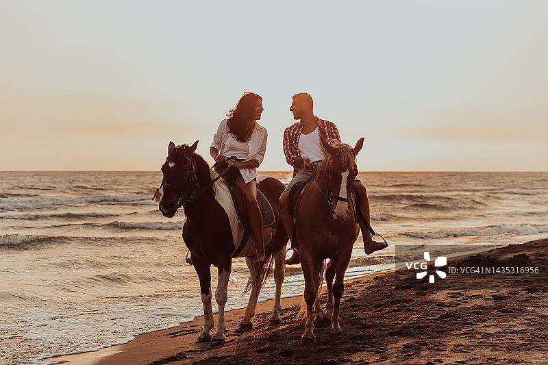 夕阳西下，一对相爱的情侣穿着夏装在沙滩上骑马。背景是大海和日落。有选择性的重点图片素材