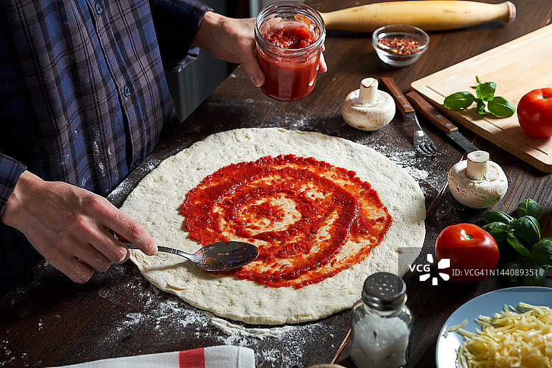 在家里，披萨店或咖啡馆用蔬菜和蘑菇烹饪意大利素食披萨。面包房的女面包师、糕点师或厨师用勺子在面团上涂番茄酱。食物的概念。一步一步的指导，自己做。步骤1。图片素材
