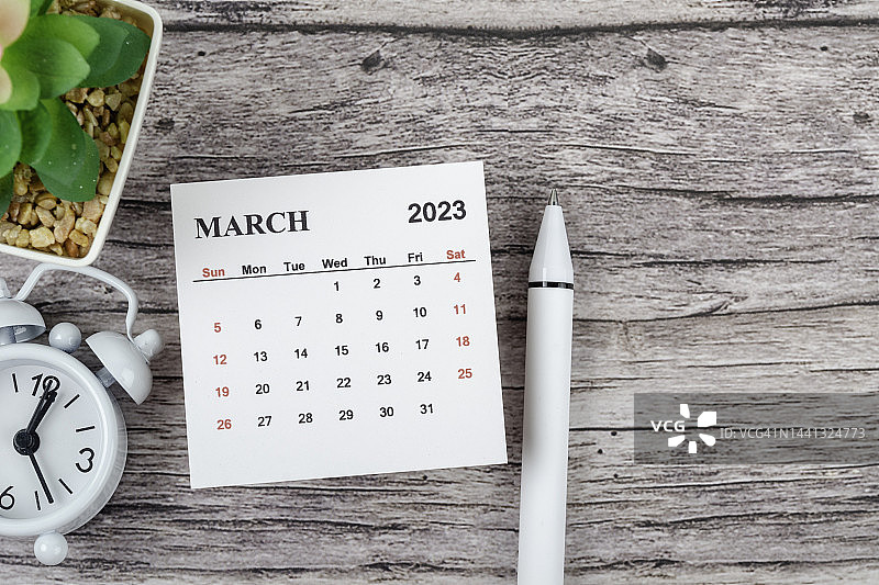 日历桌2023:3月是组织者计划和截止日期的月份，木桌背景上有一个闹钟和白笔。图片素材
