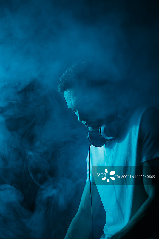 DJ在烟雾和蓝色霓虹灯的电子音乐派对上播放音乐。年轻的音乐节目主持人以夜总会为背景图片素材